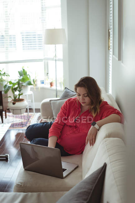 Young pregnant woman sitting on sofa using her laptop at home — Stock Photo