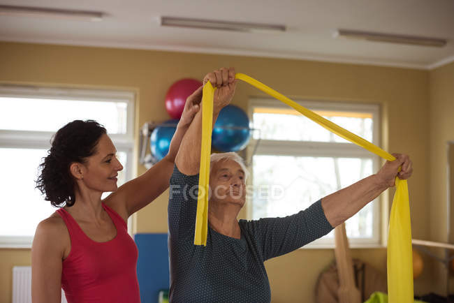 Female therapist assisting senior woman with exercise band in nursing home — Stock Photo