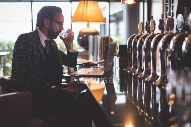 Бізнесмен використовує ноутбук, маючи віскі в барі — стокове фото