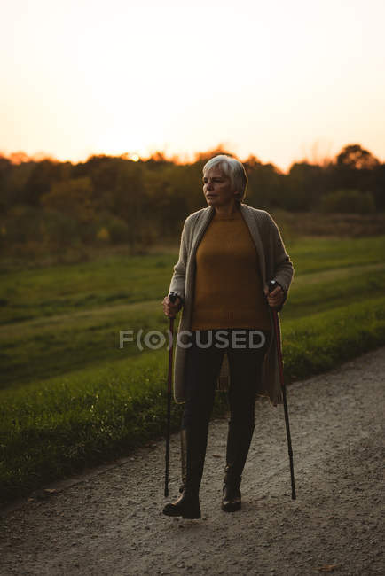 Senior woman walking on a dusty road with walking sticks at the time of dawn — Stock Photo