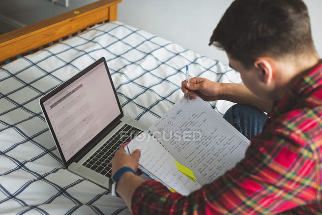 Junger Mann lernt mit Notizbuch und Laptop im Bett. — Stockfoto