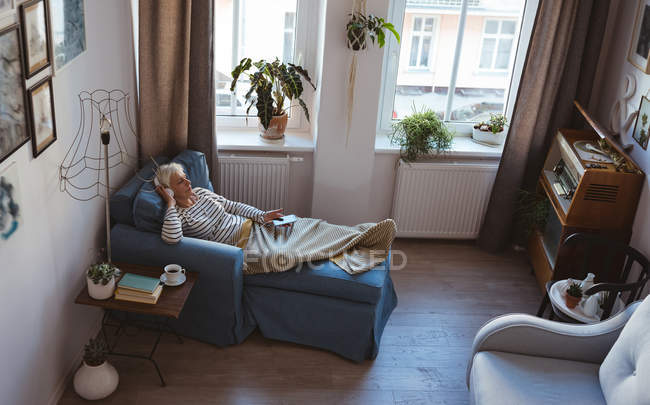 Высокоугольный вид пожилой женщины, расслабляющейся на диване со смартфоном, слушая музыку дома — стоковое фото