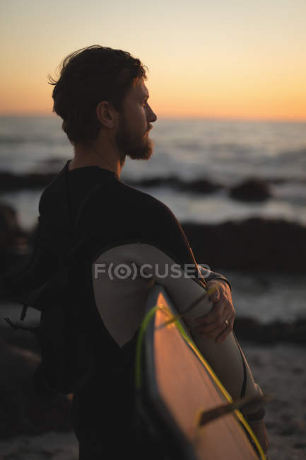 Surfista com prancha de surf em pé na praia ao pôr do sol — Fotografia de Stock
