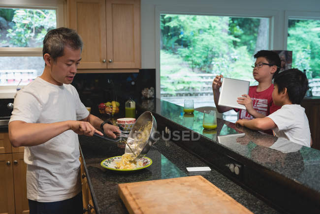 Enfants utilisant une tablette numérique pendant que le père prépare la nourriture dans la cuisine à la maison — Photo de stock