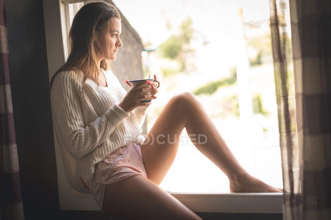 Молода жінка сидить на підвіконні, маючи каву вдома . — стокове фото