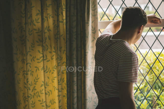 Visão traseira do homem inclinado e olhando através da janela em casa . — Fotografia de Stock