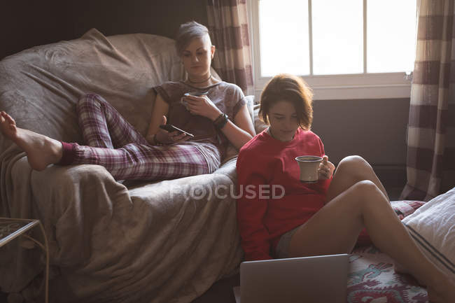 Гомосексуальна пара використовує мобільний телефон і ноутбук, маючи каву вдома . — стокове фото