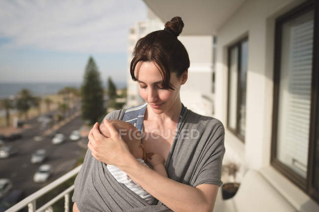 Молода мама носить дитину в слінгу на балконі в сонячний день — стокове фото
