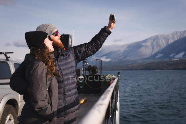 Pareja feliz tomando selfie con teléfono móvil cerca del río - foto de stock