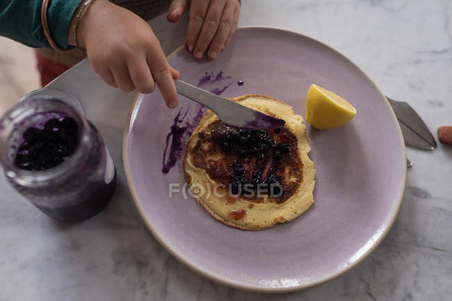 Nahaufnahme von Kinderhänden, die Marmelade auf Pfannkuchen am Tisch verteilen. — Stockfoto