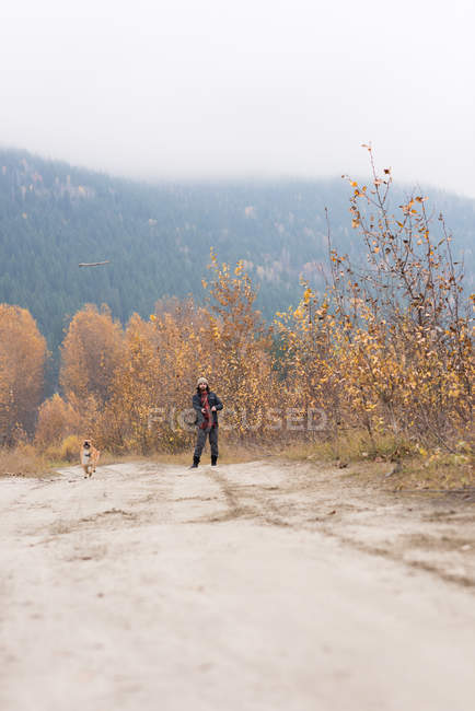 Homem e seu cão de estimação brincando no caminho vazio cercado de arbustos — Fotografia de Stock