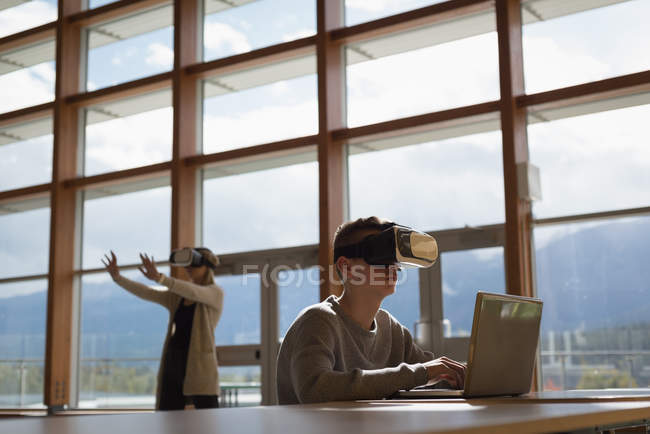 Студенты университета за столом с использованием ноутбука и гарнитуры виртуальной реальности — стоковое фото
