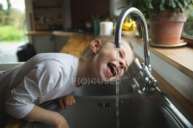 Мальчик пьет воду из крана на кухне дома — стоковое фото