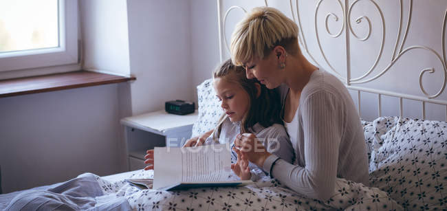 Дочь и мать читают книгу на кровати в спальне — стоковое фото
