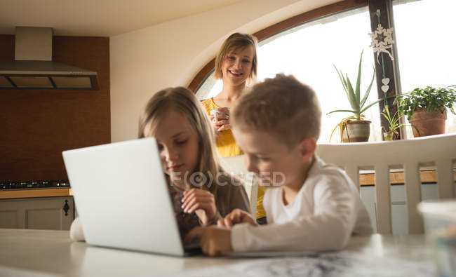 Madre guardando i bambini che utilizzano il computer portatile in cucina a casa — Foto stock