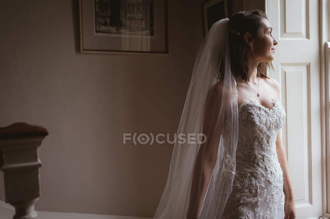 Dreamy bride looking out of the window at home — Stock Photo