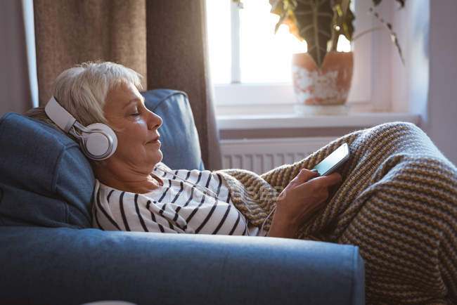 Senior woman listing to music on phone while siting on sofa in living room at home — Stock Photo