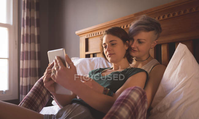 Pareja lesbiana abrazando y usando tableta digital en el dormitorio en casa . - foto de stock