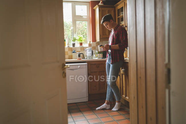 Homme utilisant un téléphone portable et tenant une tasse de café dans la cuisine à la maison . — Photo de stock