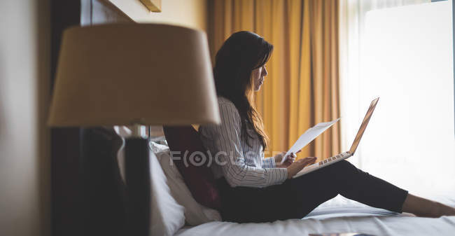 Femme d'affaires lisant des documents tout en travaillant sur ordinateur portable dans la chambre d'hôtel — Photo de stock