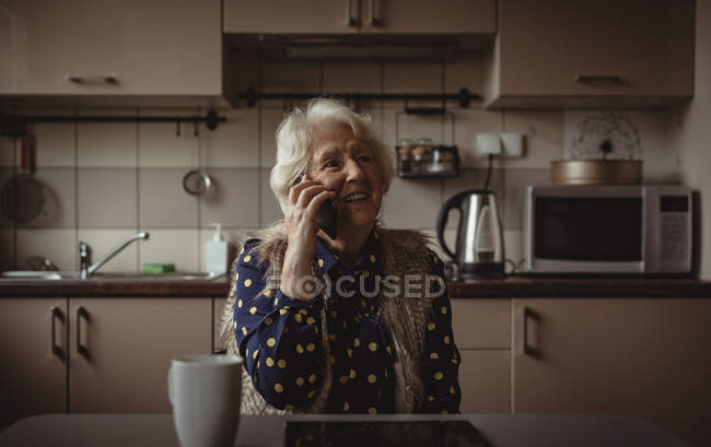 Усміхнена старша жінка розмовляє по телефону на кухні вдома — стокове фото