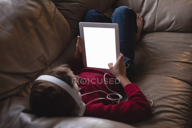 Niño usando tableta digital con auriculares en la sala de estar en casa - foto de stock