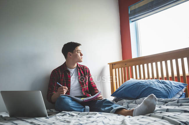Jeune homme assis sur le lit avec ordinateur portable et ordinateur portable à la maison . — Photo de stock