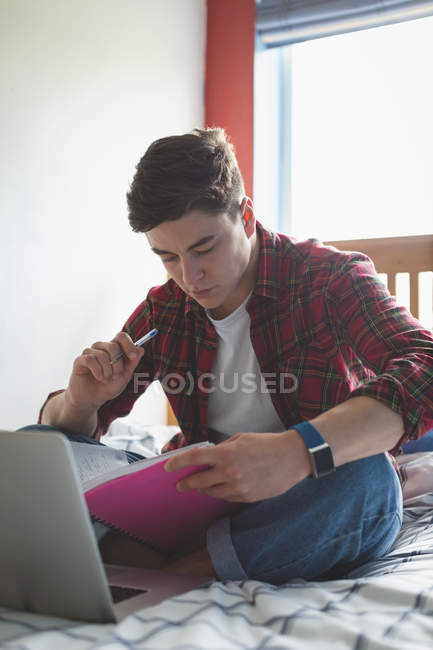 Young man working on bed with notebook and laptop at home. — Stock Photo