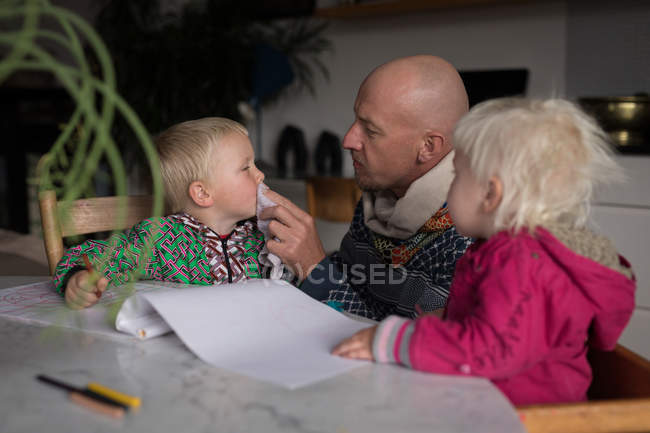 Padre limpieza hijo con servilleta mientras dibuja con los niños en la sala de estar en casa . - foto de stock