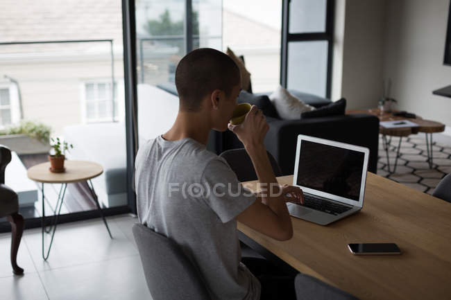 Jovem tomando café enquanto usa laptop em casa — Fotografia de Stock