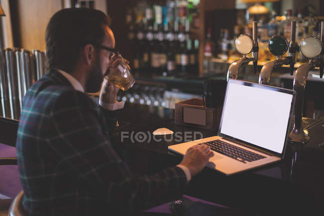 Uomo d'affari che utilizza il computer portatile pur avendo whisky nel bar — Foto stock