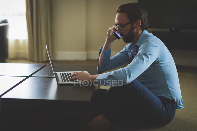 Бизнесмен разговаривает по мобильному телефону во время использования ноутбука в номере отеля — стоковое фото