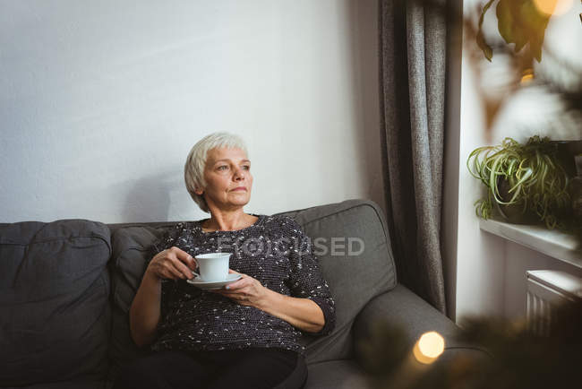Старшая женщина сидит на диване и смотрит в сторону, держа чашку кофе дома — стоковое фото