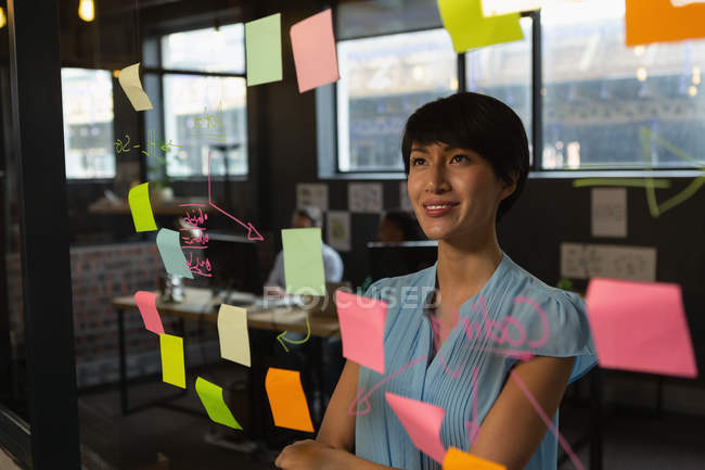Executivo feminino olhando para notas pegajosas na parede de vidro no escritório . — Fotografia de Stock