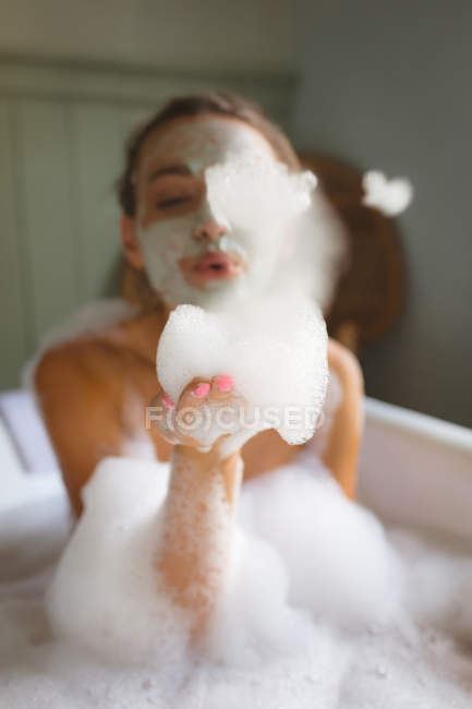 Жінка в масці для обличчя дме піну під час прийняття ванни у ванній . — стокове фото