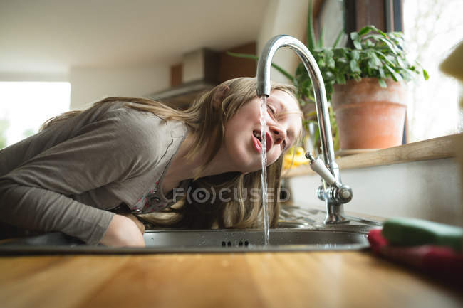 Молодая девушка пьет воду из крана на кухне дома — стоковое фото