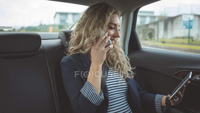 Усміхнена бізнес-леді розмовляє на мобільному телефоні, використовуючи цифровий планшет в машині — стокове фото