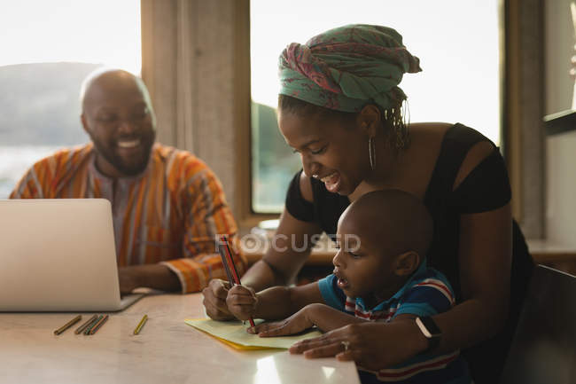Мать помогает сыну рисовать за столом с улыбающимся отцом за ноутбуком
. — стоковое фото