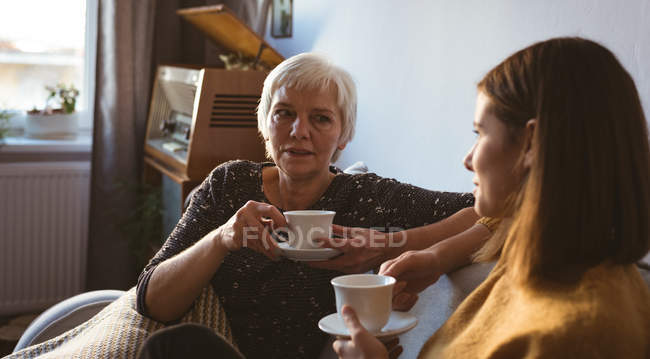 Старша жінка і дочка сидять на дивані з чашкою кави у вітальні вдома — стокове фото