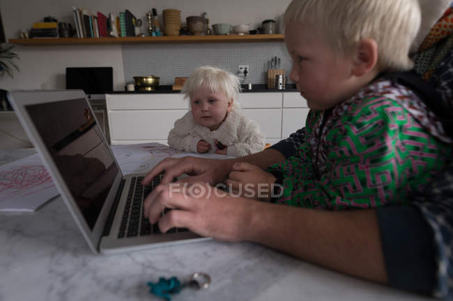 Father using laptop at table with son and daughter in kitchen at home. — Stock Photo