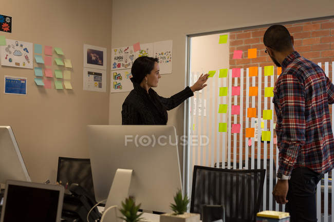 Compañeros de negocios discutiendo sobre notas adhesivas en la oficina - foto de stock