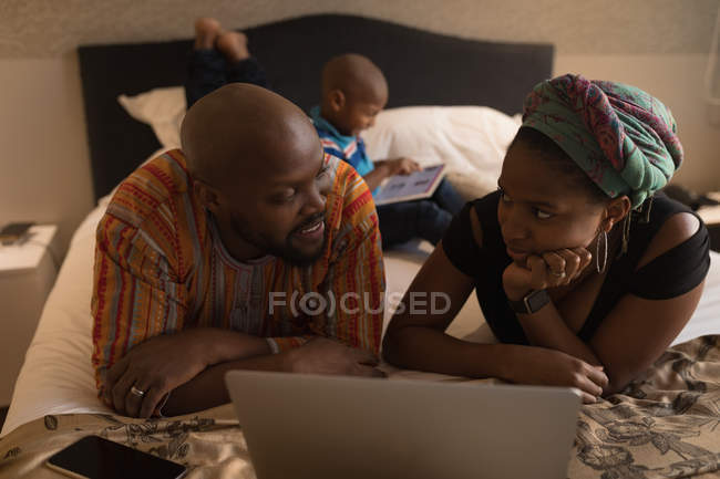 Casal usando laptop enquanto filho brincando com tablet digital no quarto em casa . — Fotografia de Stock