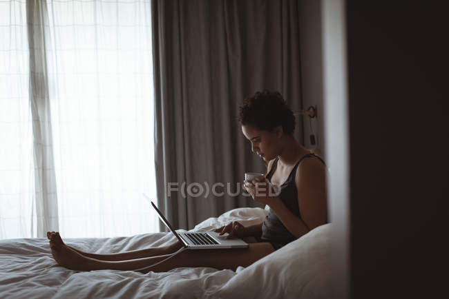 Jeune femme assise sur son lit en utilisant son ordinateur portable tout en prenant un café dans la chambre à coucher à la maison — Photo de stock