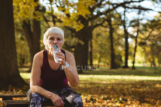 Старша жінка п'є воду в парку в сонячний день — стокове фото
