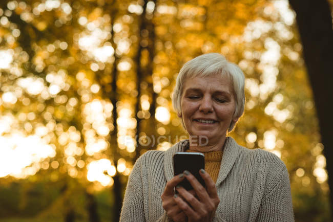 Sorrindo mulher idosa usando um telefone inteligente em um parque ao amanhecer — Fotografia de Stock