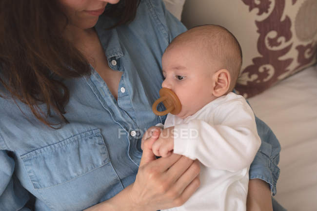 Милый ребенок с пустышкой во рту, лежащий в материнской руке дома — стоковое фото