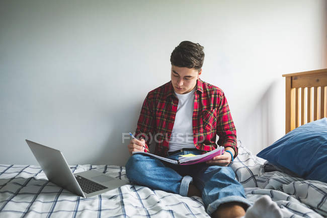 Young man studying on bed with notebook and laptop at home. — Stock Photo