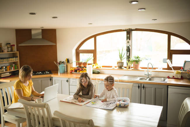Frères et sœurs dessin tandis que la mère travaillant sur ordinateur portable dans la cuisine à la maison — Photo de stock
