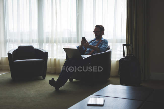 Uomo d'affari che utilizza il telefono cellulare seduto sulla poltrona in camera d'albergo — Foto stock