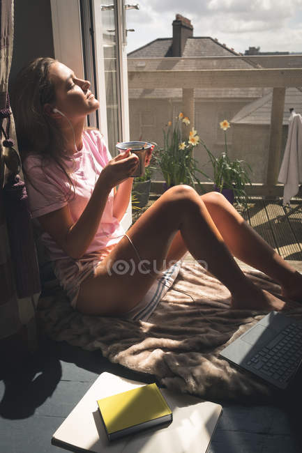 Mulher sentada perto da varanda enquanto toma café e ouve música em casa . — Fotografia de Stock